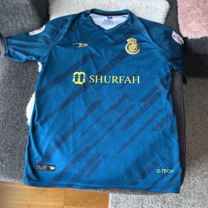 Blå fotbollströja från Al Nassr - Snygg blå fotbollströja från Al Nassr med gula detaljer och sponsorlogga. Tröjan har korta ärmar och ett mönster i olika nyanser av blått. Namnet 'Ronaldo' och nummer 7 pryder baksidan. Perfekt för fotbollsfans!  Storlek XL men passar L också