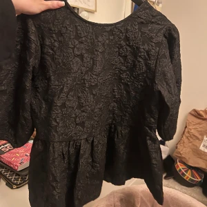 Svart blus med knytning - Elegant svart blus med texturerat mönster och knytning i ryggen. Blusen har trekvartsärmar och en lätt volangdetalj vid nederkanten. Perfekt för en stilren look.