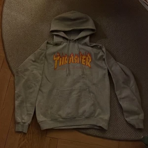 Grå hoodie från Thrasher - Säljer en grå hoodie från Thrasher med den ikoniska loggan i orange och gult på bröstet. Är i storlek S men är lite oversized, så passar M också! Priset kan diskuteras vid snabb affär. 