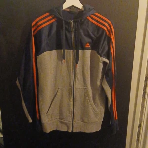 Grå och marinblå huvtröja från Adidas - Säljer en grå och marinblå huvtröja från Adidas med dragkedja och huva. Tröjan har de klassiska tre röda ränderna längs ärmarna och en ficka framtill. Perfekt för en sportig look.