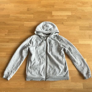 Grå hoodie från Sail Racing - Säljer en grå hoodie från Sail Racing med dragkedja framtill. Tröjan har långa ärmar och en praktisk huva. Perfekt för en avslappnad stil. 🩶