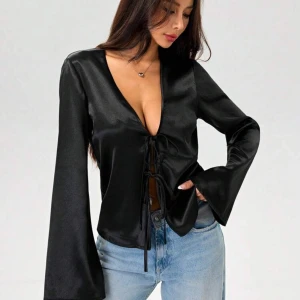 Svart satinblus med knytning - Snygg svart blus i satin med djup v-ringning och knytning framtill. Blusen har långa ärmar och en elegant, glansig finish.
