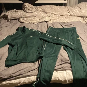 Grön träningsdräkt från Lacoste - Säljer en grön träningsdräkt från Lacoste i regular fit. Setet består av en jacka med dragkedja och matchande byxor. Båda delarna har vita detaljer längs ärmar och ben. Perfekt för en sportig look🐊