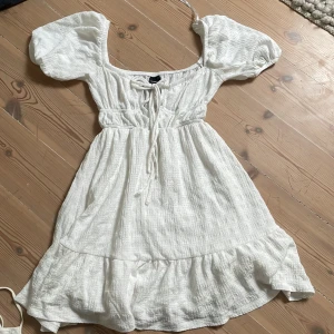Vit klänning från Gina Tricot - Charmig vit klänning från Gina Tricot med puffiga kortärmar och knytning framtill. Klänningen har en volangdetalj vid fållen och är tillverkad i ett lätt och luftigt material, perfekt för varma dagar.