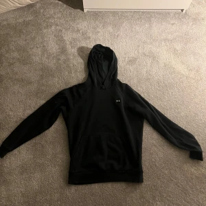 Svart hoodie från Under Armour  - Säljer en stilren svart hoodie från Under Armour. Den har en klassisk design med en liten logga på bröstet och en praktisk magficka. Perfekt för en avslappnad look eller träning. 