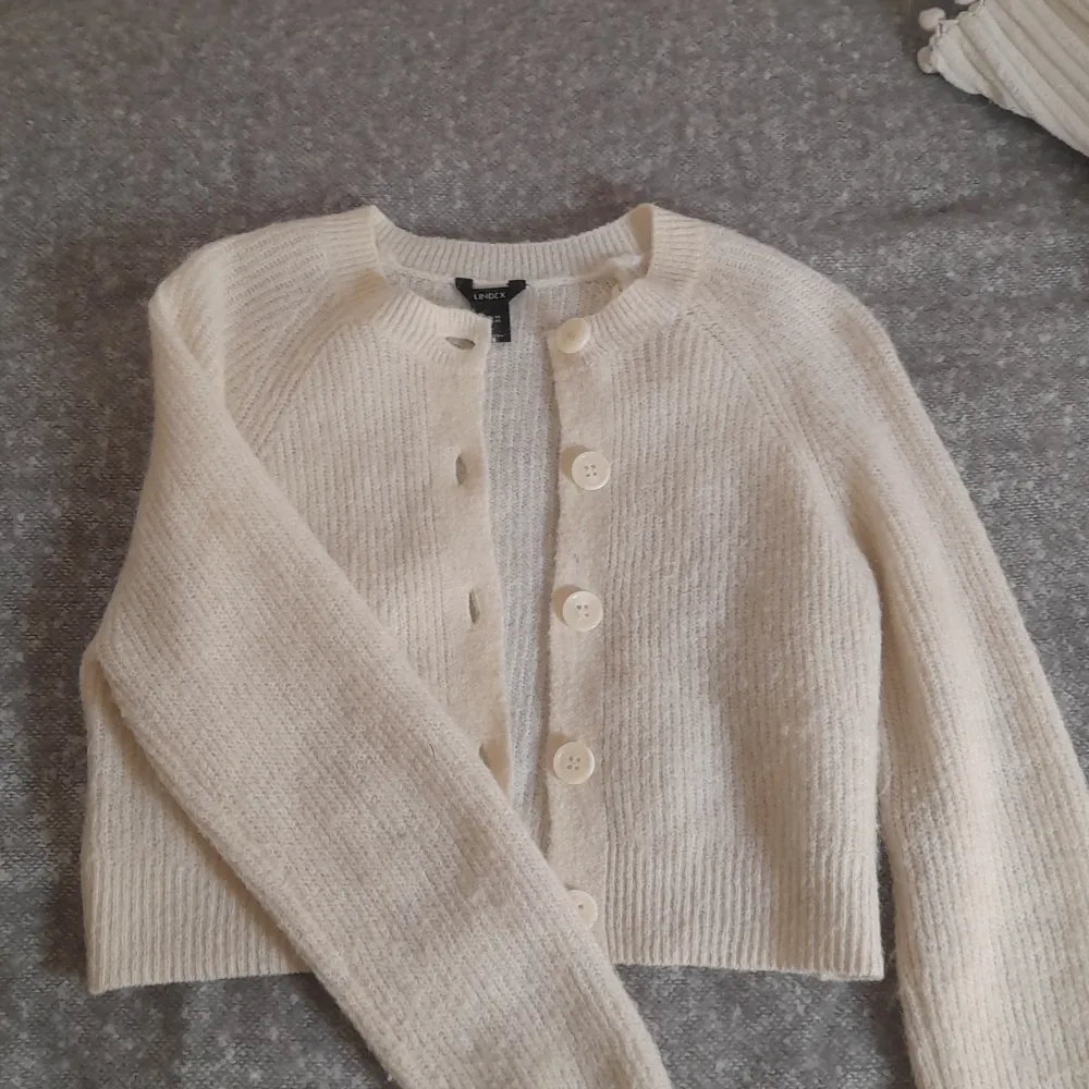 Säljer en mysig vit stickad kofta från Lindex i storlek XS. Koftan har långa ärmar och knäpps med stora knappar framtill. Perfekt för kyliga dagar och ger en stilren look.. Neuletakit & Villapaidat.