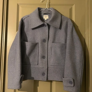 Ljusblå kappa från Kappahl - Säljer en stilren grå kappa från Kappahl i storlek XS. Jackan har en klassisk krage och knäpps med stora knappar framtill. Den har två stora fickor och långa ärmar. Perfekt för kyligare dagar!