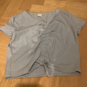 Ljusblå ribbad topp från Lindex - Säljer en ljusblå ribbad topp från Lindex med korta ärmar och knytning framtill. Toppen har en v-ringad hals och är perfekt för en avslappnad stil. Passar barn i åldern 9-12 år.