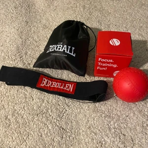 Boxbollen träningsset - Säljer ett Boxbollen träningsset som inkluderar en röd boll med elastiskt band och ett svart pannband. Perfekt för att förbättra fokus och reaktionsförmåga. Kommer med en praktisk svart förvaringspåse och en app-licens för extra träningstips! Skicket är bra det är bara använd några gånger. Orginalpris: 250kr