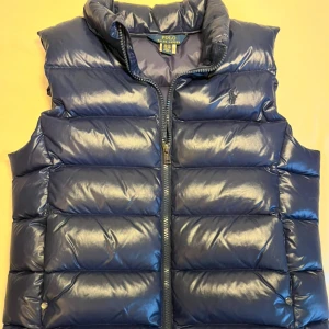 Blå dunväst från Polo Ralph Lauren - Snygg blå dunväst från Polo Ralph Lauren med dragkedja längst med västen. Västen har en quiltad design. Perfekt för kyligare dagar. Skicket på västen är 8-9/10.