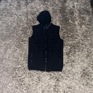 Zara väst - Snygg svart väst från Zara Man med huva och dragkedja. Perfekt för lager-på-lager stil. Västen har en stilren design och är idealisk för både vardag och mer avslappnade tillfällen.