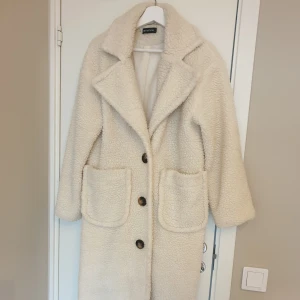 Beige teddyjacka  - Mysig beige teddykappa från PrettyLittleThings med stora knappar och två framfickor. Jackan har en klassisk krage och är perfekt för kyliga dagar. Den är långärmad och har en mjuk insida för extra komfort. *notera att den är noprig och att en innersöm gått upp, se bild.                            https://www.prettylittlething.qa/cream-maxi-borg-coat.html