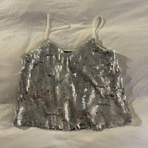 Silvrig paljettopp - Säljer en glittrig silvrig paljettopp med smala axelband. Perfekt för en festlig kväll! Toppen har en lös passform och ärmlös design.