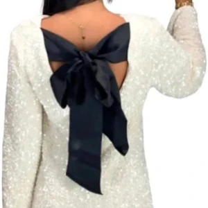 Vit glittrig klänning med svart rosett - Elegant vit klänning med glittrigt material och en stor svart rosett i ryggen. Klänningen har långa ärmar och en öppen rygg. Perfekt för festliga tillfällen. Strl M men kan passa S också😌aldrig använd då den blev försenad till nyår😊