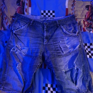 Slitna jeansshorts - Säljer ett par coola jeansshorts med slitna detaljer och en avslappnad look. Shortsen har flera fickor och en klassisk jeansblå färg. Perfekta för en casual stil.