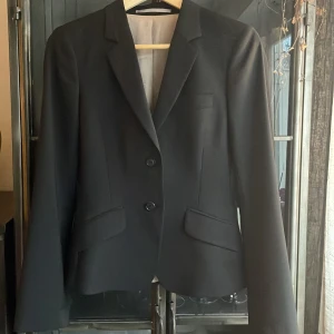 Brun kavaj från Tiger of Sweden - Elegant brun-mönstrad kavaj/blazer från Tiger of Sweden med klassisk design. Kavajen har två knappar framtill och fickor med lock. Perfekt för en stilren look! Storleken är 32, men då jag använde den var jag storlek 34/36. Den är älskad och varsamt använd, nu nyss hämtad från kemtvätten och redo att göra succé på nästa bärare!