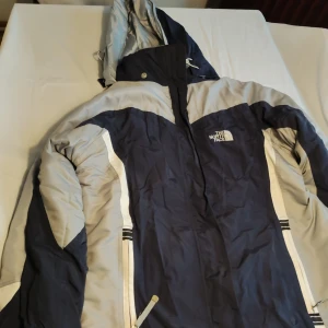 Blå och grå jacka från The North Face - Säljer en blå och grå jacka från The North Face Summit Series. Jackan har en huva och dragkedja framtill. Perfekt för kyliga dagar med sin robusta design och funktionella detaljer.