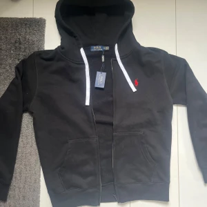 Svart hoodie från Polo Ralph Lauren - Säljer en snygg svart hoodie från Polo Ralph Lauren med dragkedja och vit snörning i huvan. Defekt dragkedja. Den har en röd broderad logotyp på bröstet och praktiska fickor framtill. Perfekt för en stilren och bekväm look.