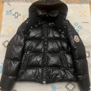 Moncler Himalaya strl 2 - Säljer en snygg svart dunjacka från Moncler med huva och dragkedja. Jackan har ett Moncler-märke på ärmen och praktiska fickor med dragkedjor. Perfekt för kyliga dagar!
