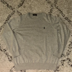 Grå tröja från Polo Ralph Lauren - Säljer en stilren grå tröja från Polo Ralph Lauren med rund hals och det klassiska logotypbroderiet på bröstet. Tröjan har långa ärmar och ribbade muddar vid ärmslut och nederkant. Perfekt för en avslappnad och snygg look.