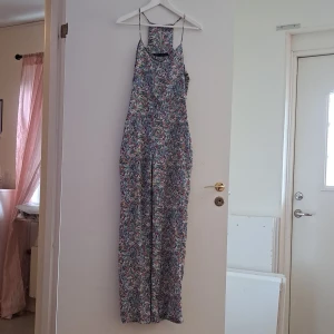 Blommig flerfärgard jumpsuit - Säljer en superfin blommig jumpsuit med smala axelband. Perfekt för sommardagar eller en avslappnad fest. Det har en lös passform och är gjord i ett lätt och bekvämt material. Den har ett färgglatt mönster som verkligen sticker ut!
