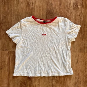 Söt vit t-shirt med röd krage från H&M - En vit t-shirt från H&M:s Divided-kollektion. Den har en röd krage och texten 'LOVE' tryckt på framsidan. T-shirten är i storlek XS och är gjord av 100% bomull, vilket gör den superbekväm och perfekt för vardagsbruk.