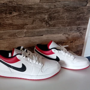 Nike sneakers i vitt, svart och rött - Säljer ett par snygga Nike sneakers i vitt med svarta och röda detaljer. Skorna har en klassisk design med snörning och en röd sula som ger en cool kontrast. Perfekta för både vardag och fest!