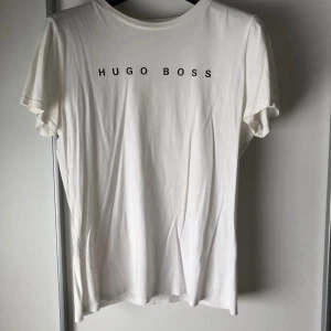 Vit t-shirt från Hugo Boss - Säljer en vit t-shirt från Hugo Boss. Den har korta ärmar. Perfekt för en avslappnad och snygg look. Hugo Boss-loggan är tryckt både fram och bak på tröjan. Knappt använd