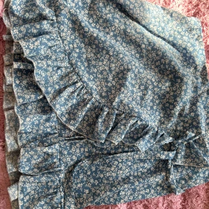 Blommig volangkjol i blått och vitt - Säljer en superfin blommig kjol i blått med vita detaljer. Kjolen har volanger som ger den en extra söt touch. Perfekt för sommarens alla fester eller en mysig dag på stan. Materialet känns lätt och luftigt, vilket gör den idealisk för varma dagar.