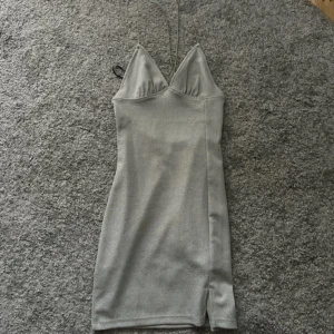 Grå klänning från H&M Divided - Säljer en snygg silvrig klänning från H&M. Den är ärmlös med smala axelband och en öppen rygg med korsade band. Klänningen har en liten slits på sidan och är i ett glittrigt material som passar perfekt för fest. Den är i storlek XS och sitter tight för en smickrande passform. Den är helt oanvänd, endast testad.