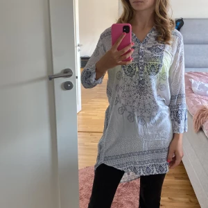 Vit och blå tunika med mönster - Säljer en luftig och bekväm vit tunika med blått mönster. Den har trekvartsärmar och en lös passform, perfekt för vår och sommar. Tunikan har en v-ringad hals och är längre baktill vilket ger en snygg siluett.