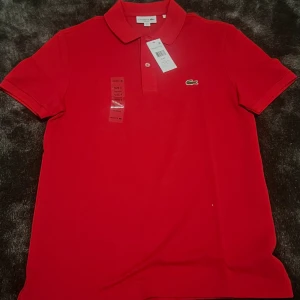 Röd pikétröja från Lacoste - Säljer en snygg helt oanvänd röd pikétröja från Lacoste. Den har en klassisk krage med två knappar och det ikoniska krokodilmärket på bröstet. Perfekt för en stilren och avslappnad look.