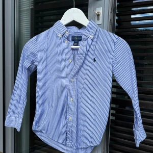  Ralph Lauren skjorta i nyskick  6 år  - Vit och blå randig Ralph Lauren skjorta med blå häst. Button down krage. Mycket fint skick, knappt använt.