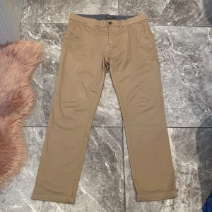 Beige chinos från Dressmann - Säljer ett par snygga beige chinos från Dressmann. De har en klassisk design med knapp och dragkedja i gylfen. Perfekta för både vardag och lite mer uppklädda tillfällen. Storleken är 33/32, vilket passar de flesta. Materialet känns slitstarkt och bekvämt.