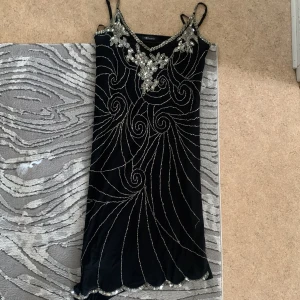 Svart klänning från Envie - Säljer en elegant svart klänning från Envie. Den har tunna axelband och är dekorerad med glittriga detaljer i ett virvlande mönster. Perfekt för en festlig kväll!