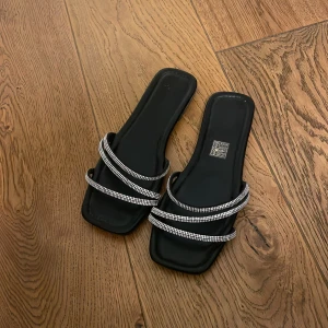 Svarta sandaler med glittriga detaljer - Snygga svarta sandaler med fyrkantig tå och dubbla remmar som är täckta med glittriga paljetter. Perfekta för en festlig kväll eller en somrig dag när du vill glänsa lite extra. De är både stilrena och bekväma!