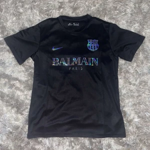 Svart fotbollströja från Nike och Balmain - Säljer en snygg svart fotbollströja i samarbete mellan Nike och Balmain. Tröjan har korta ärmar och ett stilrent tryck med Balmain Paris och en klubbemblem på bröstet. Perfekt för både träning och vardag!