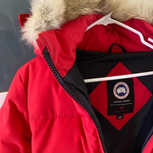 Röd dunjacka från Canada Goose - Säljer en supervarm och snygg röd dunjacka från Canada Goose. Jackan har en fluffig pälsfodrad huva och är perfekt för kalla vinterdagar. Jackan är i bra skick och redo för nya äventyr!
