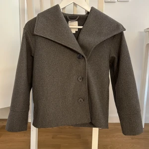 Grå kappa från Gina Tricot - Säljer en stilren grå kappa från Gina Tricot. Den har en snygg, bred krage och knäpps med tre stora knappar framtill. Perfekt för höst och vår, och ger en elegant touch till vilken outfit som helst. Kappan är i bra skick och redo för en ny ägare!