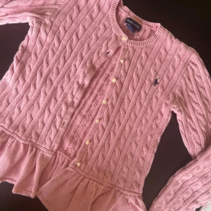 Rosa stickad kofta från Ralph Lauren - Säljer en superfin rosa stickad kofta från Ralph Lauren. Den har ett klassiskt flätmönster och knäpps med söta vita knappar. Koftan har långa ärmar och en volangdetalj vid nederkanten som ger den en extra touch av stil. Perfekt för både höst och vår! 