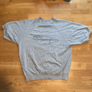 Heavyweight cropped t-shirt från Champion - Säljer en grå heavyweight cropped tröja från Champion i storlek Large. Den är gjord av 65% bomull och 35% akryl, vilket gör den både bekväm och hållbar. Perfekt för en avslappnad stil med sitt klassiska Champion-tryck på framsidan.