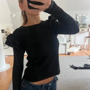 Svart ribbad tröja med volangdetaljer - Säljer en superfin svart ribbad tröja med volangdetaljer på axlarna. Den är långärmad och har en tight passform som framhäver figuren. Perfekt för både vardag och fest! 🖤