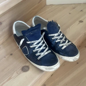 Blå sneakers från Philippe Model - Säljer ett par snygga blå sneakers från Philippe Model. Skorna har en cool design med vita snören som har text på sig. De är tillverkade i skinn och har en vit sula som ger en skön kontrast till det blå. Perfekta för en avslappnad stil!