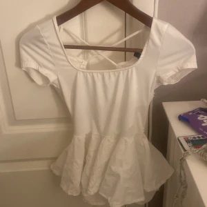 Vit topp med volangdetaljer - Säljer en superfin vit topp med volangdetaljer och korta ärmar. Toppen har en söt öppen rygg med korsade band som ger en extra touch. Perfekt för sommardagar eller en festlig kväll!