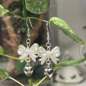 Söta örhängen med rosett och hjärta - Superfina handgjorda örhängen med en vit pärlemorliknande rosett och ett hängande silverfärgat hjärta. Perfekta för att ge en gullig touch till vilken outfit som helst.