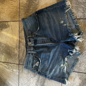 Blå jeansshorts från Levi's 501 - Säljer ett par klassiska 501 blå jeansshorts från Levi's med fransiga kanter och femficksdesign. Dessa shorts är perfekta för en avslappnad stil och har en knappgylf. Levi's-loggan syns tydligt på baksidan. Perfekta för sommardagar! 🌞