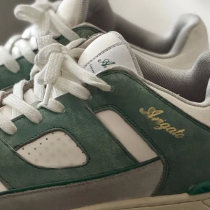 Gröna och vita sneakers från Axel Arigato - Snygga sneakers från Axel Arigato i grönt och vitt med grå detaljer. Skorna har en klassisk snörning och är tillverkade i en kombination av skinn och syntetmaterial. Perfekta för en stilren look.