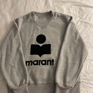 Grå sweatshirt från Isabel Marant - Säljer en stilren grå sweatshirt från Isabel Marant med ett svart tryck framtill. Tröjan har en rund halsringning och ribbade muddar vid ärmslut och nederkant. Perfekt för en avslappnad look.