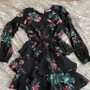 Blommig klänning med volanger - Säljer en söt blommig klänning från Ü med volanger och långärmad design. Klänningen har en elastisk midja och ett vackert blommönster i rosa och grönt på en svart bakgrund. Perfekt för en stilfull look!