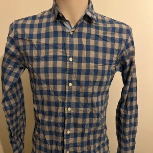 Blå och vit rutig skjorta från Jack & Jones - Snygg blå och vit rutig skjorta från Jack & Jones. Skjortan har långa ärmar och knappar framtill. Perfekt för en stilren look med en klassisk krage.